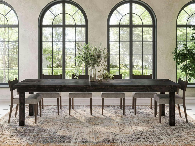 A modern dining room showcases the Farmhouse Dining Table - Charred Ember by James+James, paired with eight gray cushioned chairs. Large arched windows flood the room with natural light, while a lush green foliage centerpiece adorns the table, complemented by a patterned rug and a potted plant.