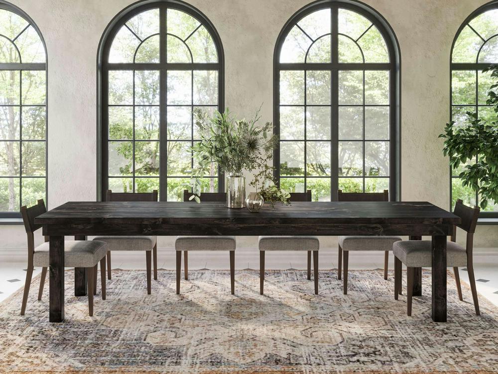 A modern dining room showcases the Farmhouse Dining Table - Charred Ember by James+James, paired with eight gray cushioned chairs. Large arched windows flood the room with natural light, while a lush green foliage centerpiece adorns the table, complemented by a patterned rug and a potted plant.