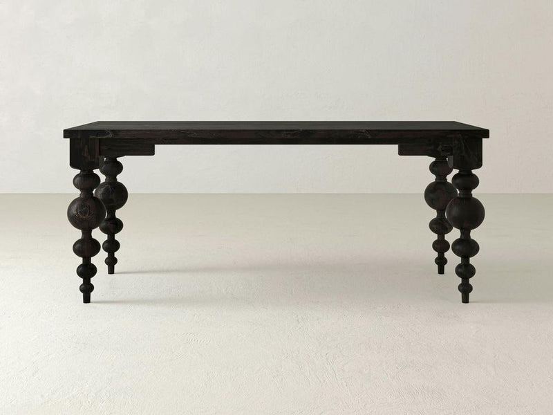 In a minimalist, light-colored room, an Olivia Desk - Charred Ember by James+James stands alone. This black, rectangular wooden desk boasts a smooth top and four intricately carved baluster-style legs that add a touch of elegance to its simple design.