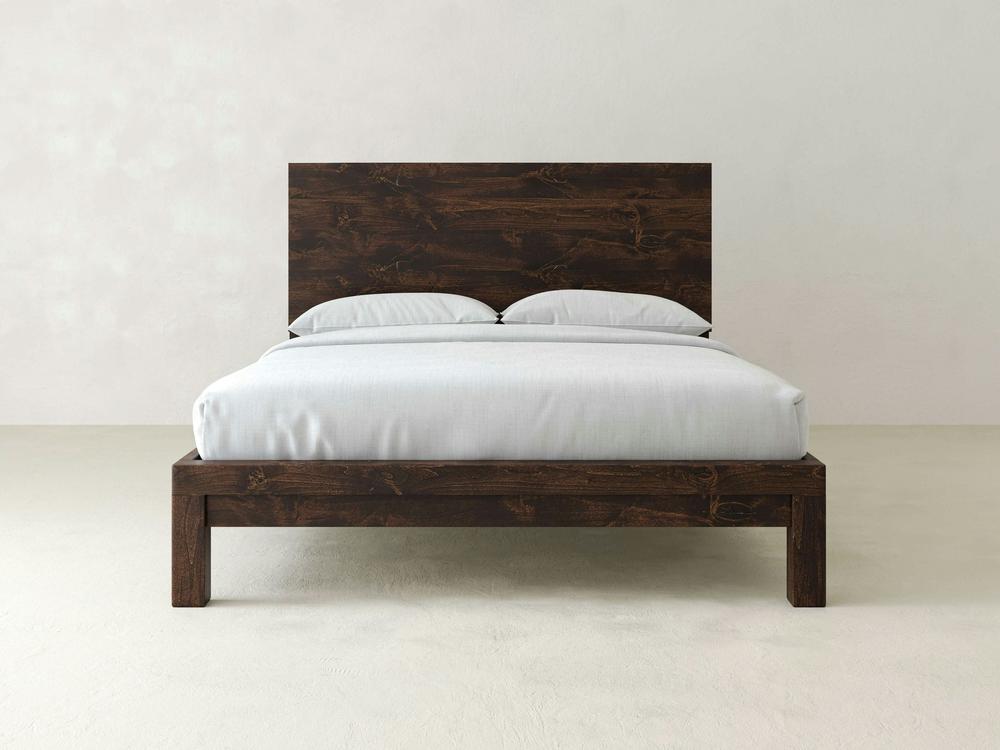 The James+James Sawyer Bed - Tobacco on Alder features a dark wood frame and headboard, paired with a white mattress, bedsheets, and two pillows, set against a plain light-colored wall and floor.