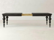 The Bailey Expandable Dining Table - Charred Ember from James+James is shown in its extended form. The table features dark-colored turned legs and is split down the middle, displaying a wooden extension mechanism. The background consists of a plain off-white wall and floor.