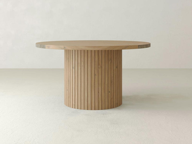 A round wooden Arthur Dining Table by James+James features a smooth, flat top and a cylindrical base adorned with vertical slats. Showcasing traditional craftsmanship, the table is set in a minimalist environment with a light, neutral-colored background.