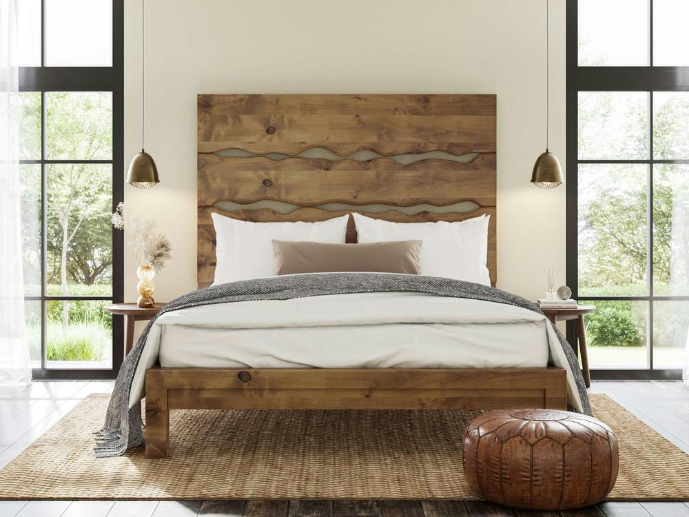 A cozy bedroom features the James+James Live Edge Bed - Harvest Wheat on Alder, topped with white bedding and a gray blanket. Pendant lights, side tables, a woven rug, brown leather pouf, and large windows complete the look.