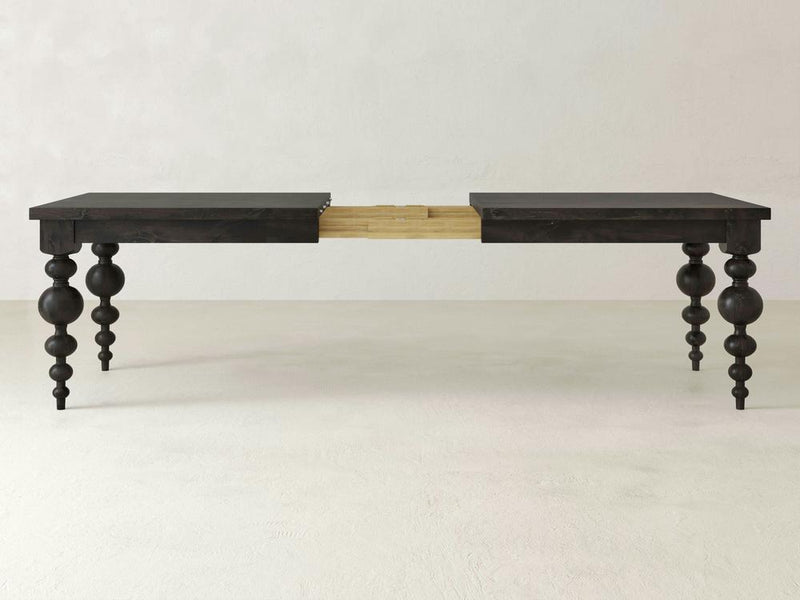 The Olivia Expandable Dining Table - Charred Ember by James+James features a dark brown wooden finish with intricately carved, turned legs. The table is extended in the center, highlighting its extension mechanism, which allows it to be lengthened. The background showcases a plain, light-colored wall and floor.