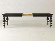 The Olivia Expandable Dining Table - Charred Ember by James+James features a dark brown wooden finish with intricately carved, turned legs. The table is extended in the center, highlighting its extension mechanism, which allows it to be lengthened. The background showcases a plain, light-colored wall and floor.