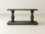 The Vivien Sofa Table by James+James is a rustic wooden console featuring a dark finish that seamlessly blends form and function. Its design includes a flat top supported by two ornately carved, baluster-style legs connected to a thick rectangular base, with its robust construction standing out against the simple, neutral background.