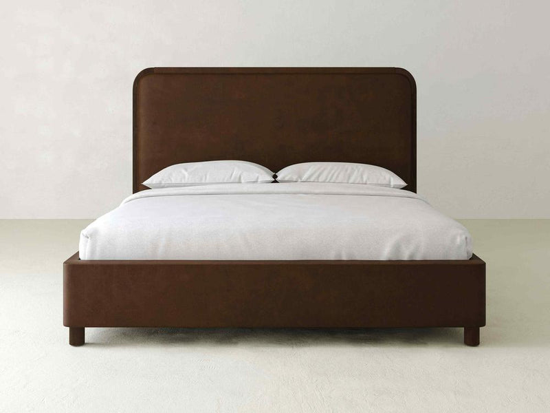 The Sloane Bed – Leather by James & James showcases modern elegance with its dark brown wooden frame and rectangular upholstered headboard. A neatly made bed with white sheets and two pillows stands out against a plain, light-colored wall.