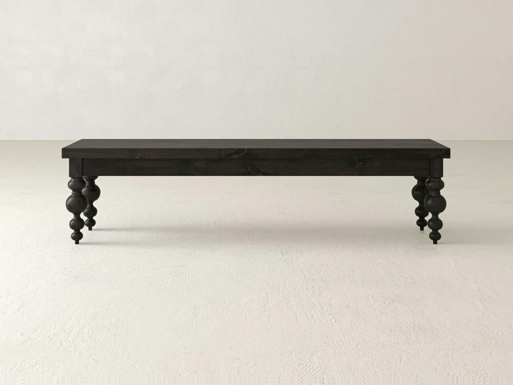 The Olivia Bench - Charred Ember on Alder by James+James, featuring ornate turned legs, is positioned against a plain, light-colored wall and floor.