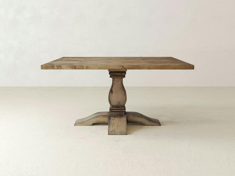A Heirloom Square Dining Table by James+James, featuring a rectangular top and a single ornate pedestal base, stands against a plain beige background. The timeless craftsmanship of the natural wood grain and rustic finish imparts an elegant, vintage appearance to the table.