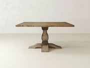 A Heirloom Square Dining Table by James+James, featuring a rectangular top and a single ornate pedestal base, stands against a plain beige background. The timeless craftsmanship of the natural wood grain and rustic finish imparts an elegant, vintage appearance to the table.