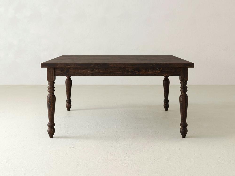 A French Country Square Dining Table in Tobacco finish from James+James stands on a light-colored floor against a plain, light-colored wall. The table features ornate, carved legs and a square top with a classic design, giving it a vintage or rustic appearance.