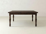 A French Country Square Dining Table in Tobacco finish from James+James stands on a light-colored floor against a plain, light-colored wall. The table features ornate, carved legs and a square top with a classic design, giving it a vintage or rustic appearance.