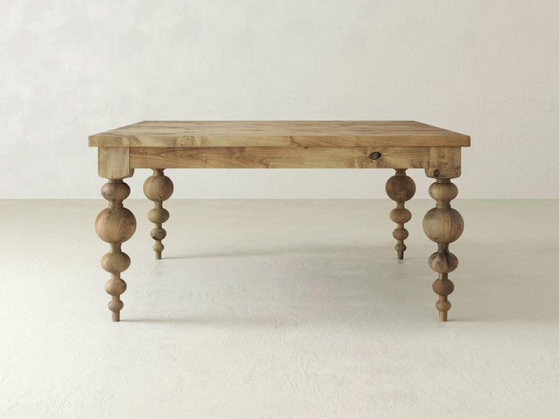A close-up of the Olivia Square Dining Table in Harvest Wheat by James+James reveals a wooden surface with a light brown color and visible grain patterns. The texture showcases irregular lines and a small knot on the right side. The surface appears smooth with natural variations in tones and shades.