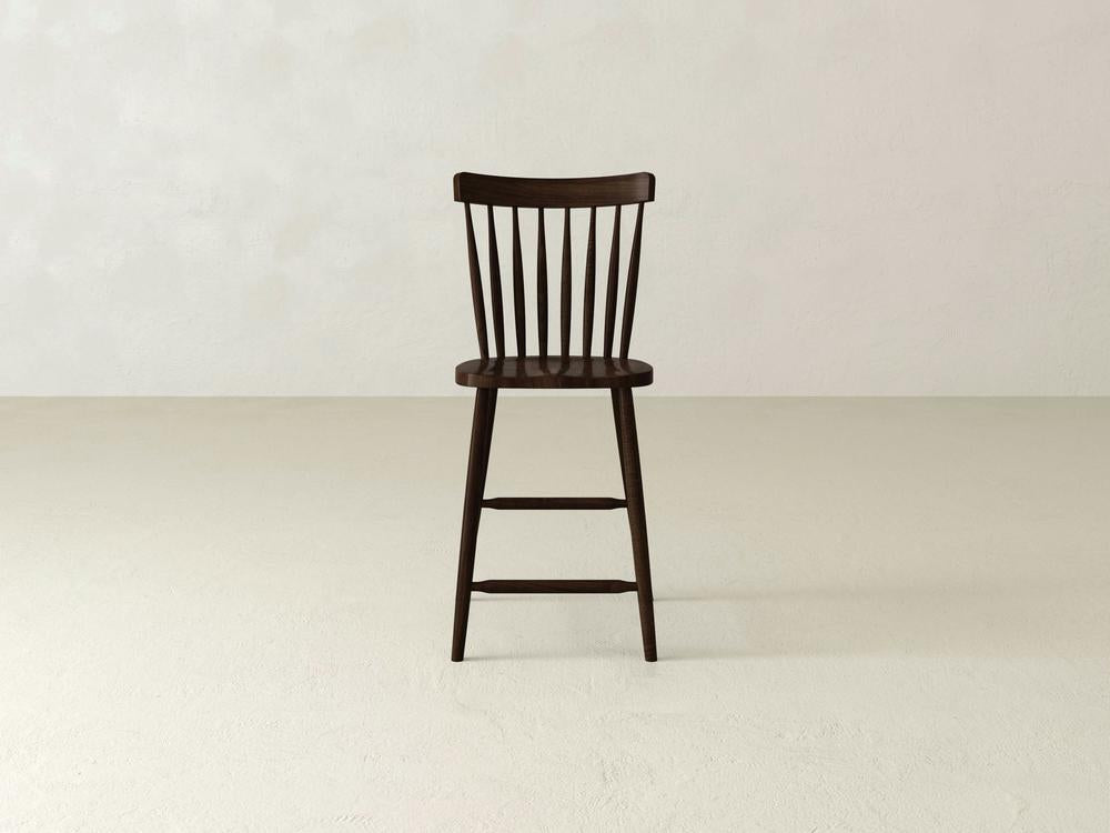 A Rustic Windsor Stool by James & James, made of durable maple hardwood, boasts a dark wooden spindle backrest. It stands on a light-colored floor in an empty room with textured white walls.