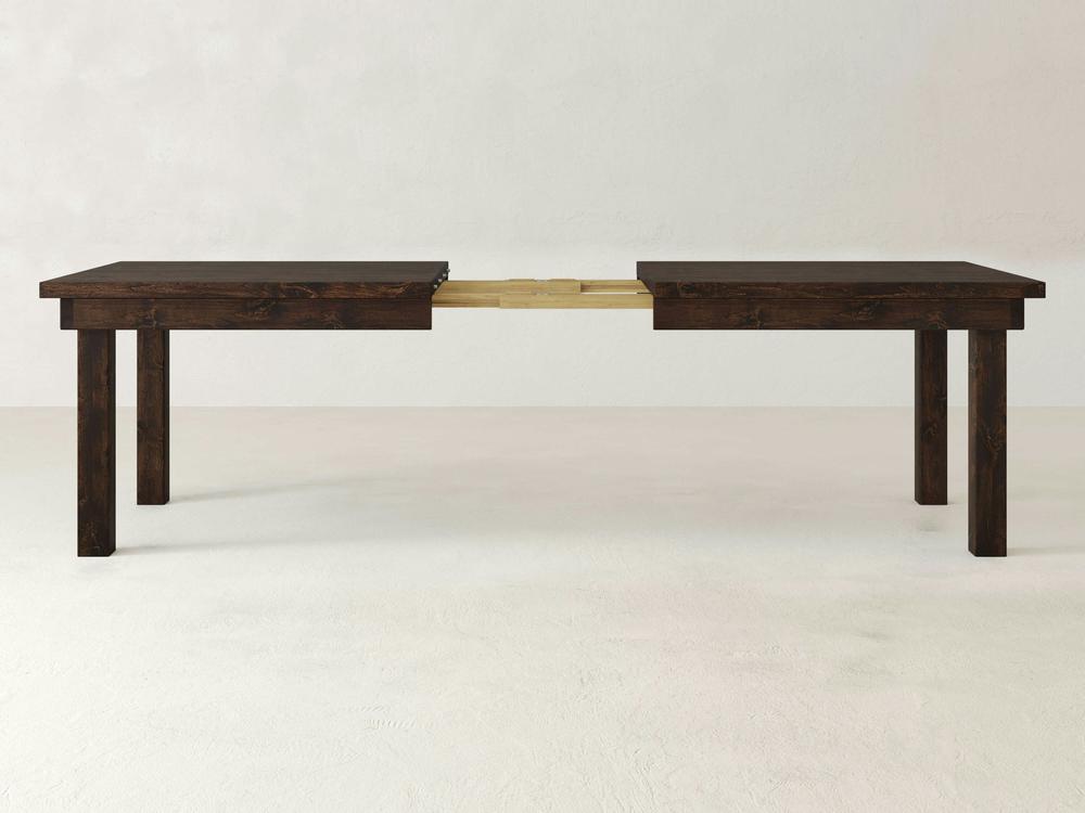 A James+James Farmhouse Expandable Dining Table, exuding rustic American elegance, is displayed against a plain white background. The dark wooden table with straight legs is partially extended in the middle, showcasing its extension mechanism.