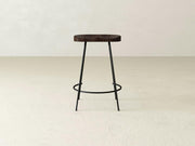 The Bryer Stool by James & James offers rustic charm with a minimalist wooden design, featuring a dark round seat and thin black metal legs. Its circular footrest adds an industrial touch, especially on light floors against off-white walls.