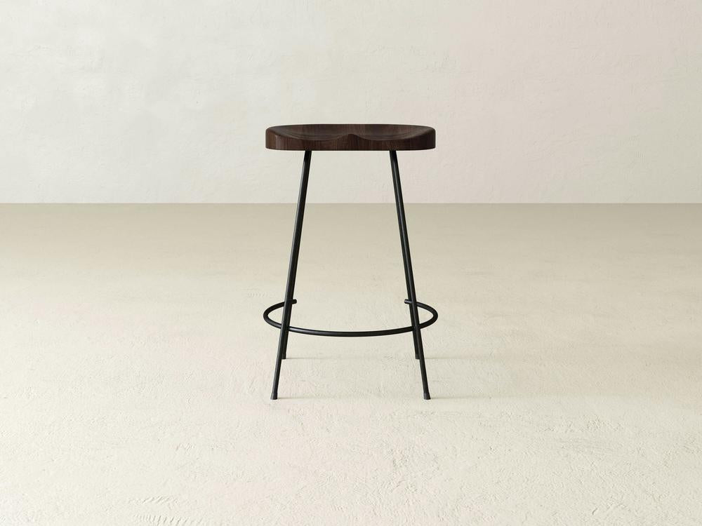 The Bryer Stool by James & James offers rustic charm with a minimalist wooden design, featuring a dark round seat and thin black metal legs. Its circular footrest adds an industrial touch, especially on light floors against off-white walls.