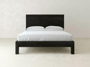 The Sawyer Bed - Charred Ember on Alder by James+James features a minimalist black wooden frame with a tall headboard and white bedding, including two pillows, set against an off-white wall and floor.
