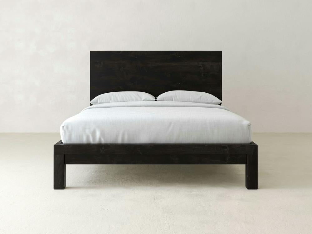 The Sawyer Bed - Charred Ember on Alder by James+James features a minimalist black wooden frame with a tall headboard and white bedding, including two pillows, set against an off-white wall and floor.