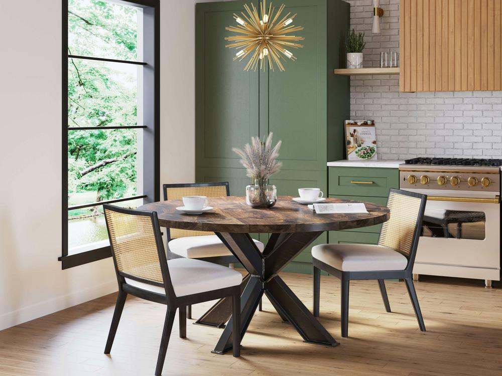 A modern dining area showcases the Shiloh Round Dining Table in Tobacco by James+James, with a cross-legged metal base, surrounded by four cushioned chairs featuring wicker backs. A starburst chandelier hangs above. Green cabinets and stainless steel appliances enhance the kitchen in the background.