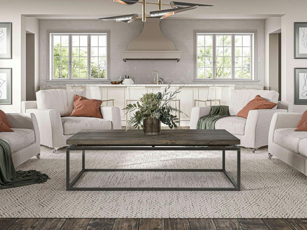 A modern living room features four light-colored armchairs with earthy-toned pillows and throws set around a Floating Top Coffee Table - Deep Grey by James+James on a textured rug. The room has two large windows letting in natural light, a minimalist kitchen in the background, and framed art on the walls.