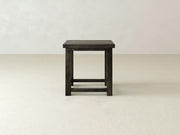 The Carter X Side Table by James+James, composed of dark hardwood, stands on a bare, light-colored floor against a plain white wall. With its minimalist design, the table showcases a rectangular top supported by four straight legs connected with horizontal supports near the base. This elegant and modern piece is ideal for any living room setting.