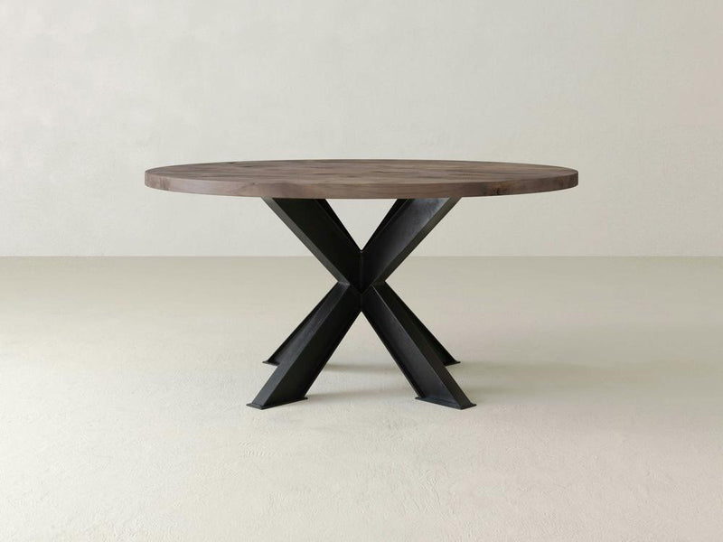 The James+James Shiloh Round Dining Table boasts a dark brown solid hardwood tabletop and a modern, crossed industrial steel base standing on a light-colored floor. Its minimalist design features clean lines and blends natural and industrial elements seamlessly.