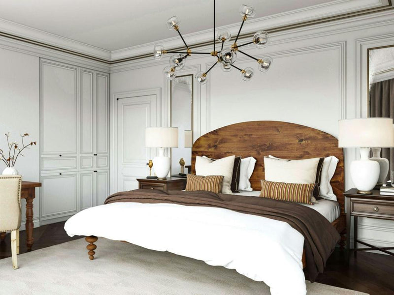 A chic bedroom featuring the Olivia Arch Bed by James+James with white bedding and brown blankets. Two bedside tables hold white lamps, a modern chandelier hangs above, and beige walls feature molding. A desk with a chair and a vase of branches completes the look.