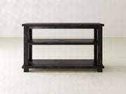 The Carter X Sofa Table in Charred Ember by James+James is a dark wooden console table with three tiers, standing on a light-colored floor against a light background. It features a simple, rectangular design with a sturdy base and open shelving for storage or display.