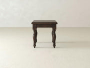 A single James+James Abigail End Table crafted from dark wood, showcasing a sophisticated turned leg motif, stands on a light beige floor against a plain, off-white wall. This rectangular table exemplifies superior craftsmanship and features detailed carvings on its legs. The setting is minimalistic and clean.