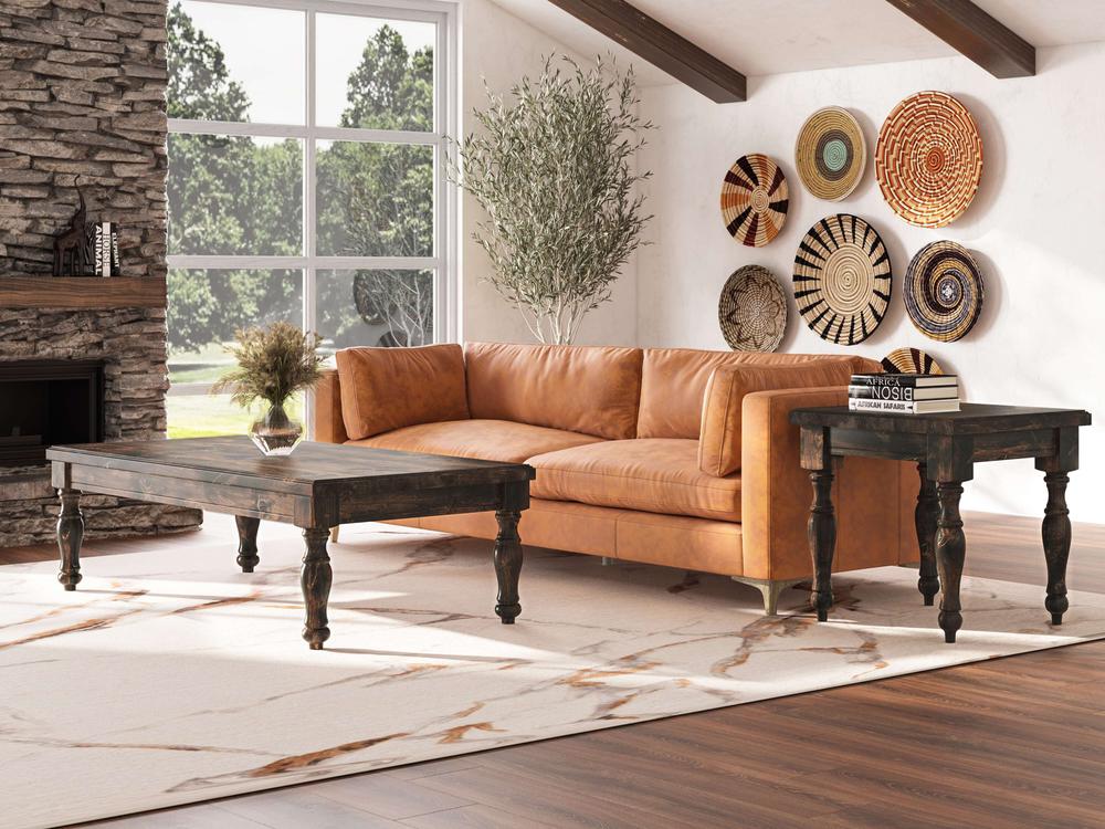 A stylish living room features a tan leather couch, two Abigail End Tables in Charred Ember by James+James, and a large potted plant. Above the couch, woven baskets of various patterns adorn the wall. A large window lets in natural light, illuminating the stone fireplace and wooden beams.