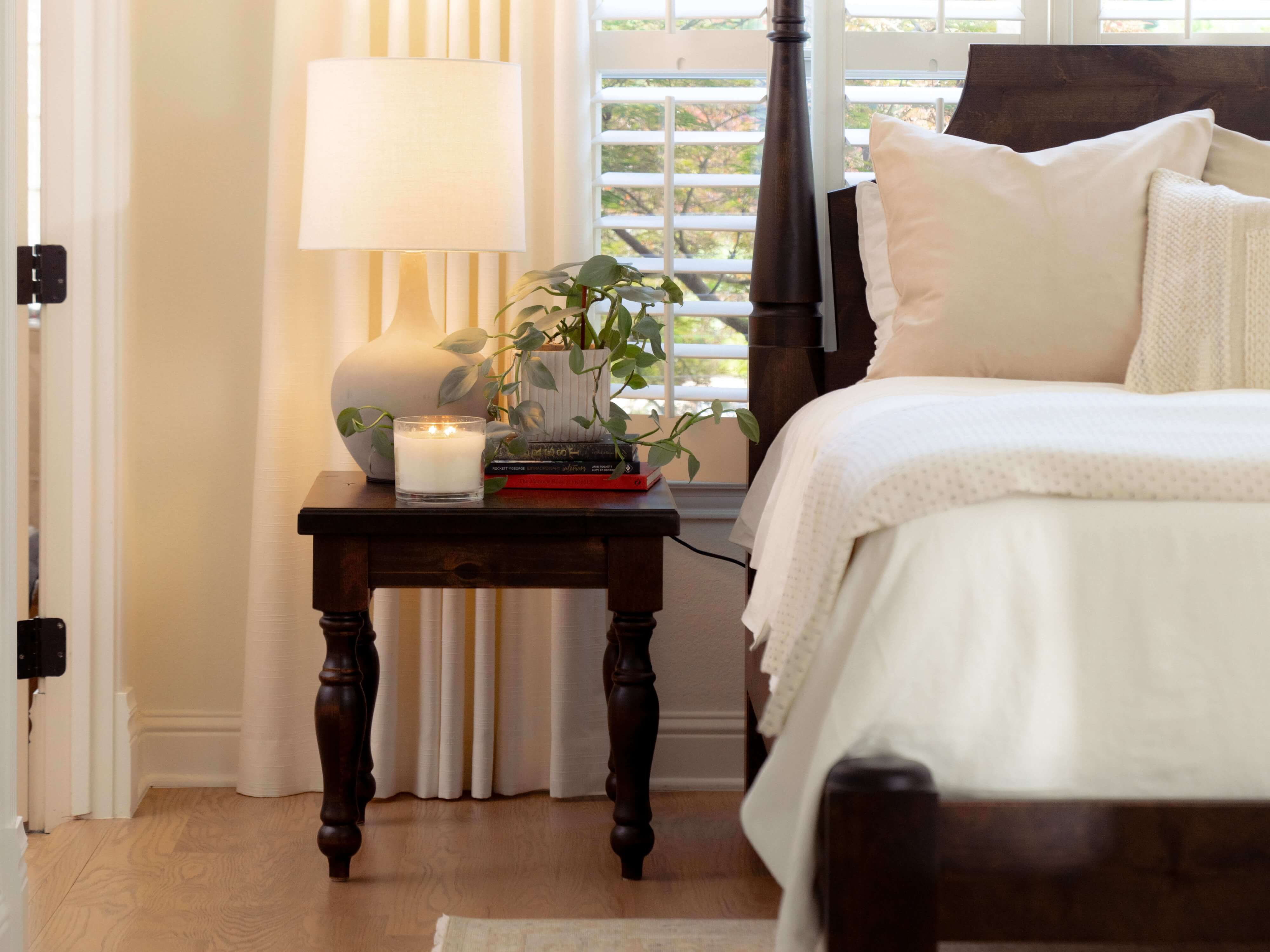 Cozy bedroom corner with a wooden nightstand holding a lamp, a lit candle, and a potted plant. A bed with white bedding is beside it, and sunlight filters through plantation shutters, casting soft light across the room.