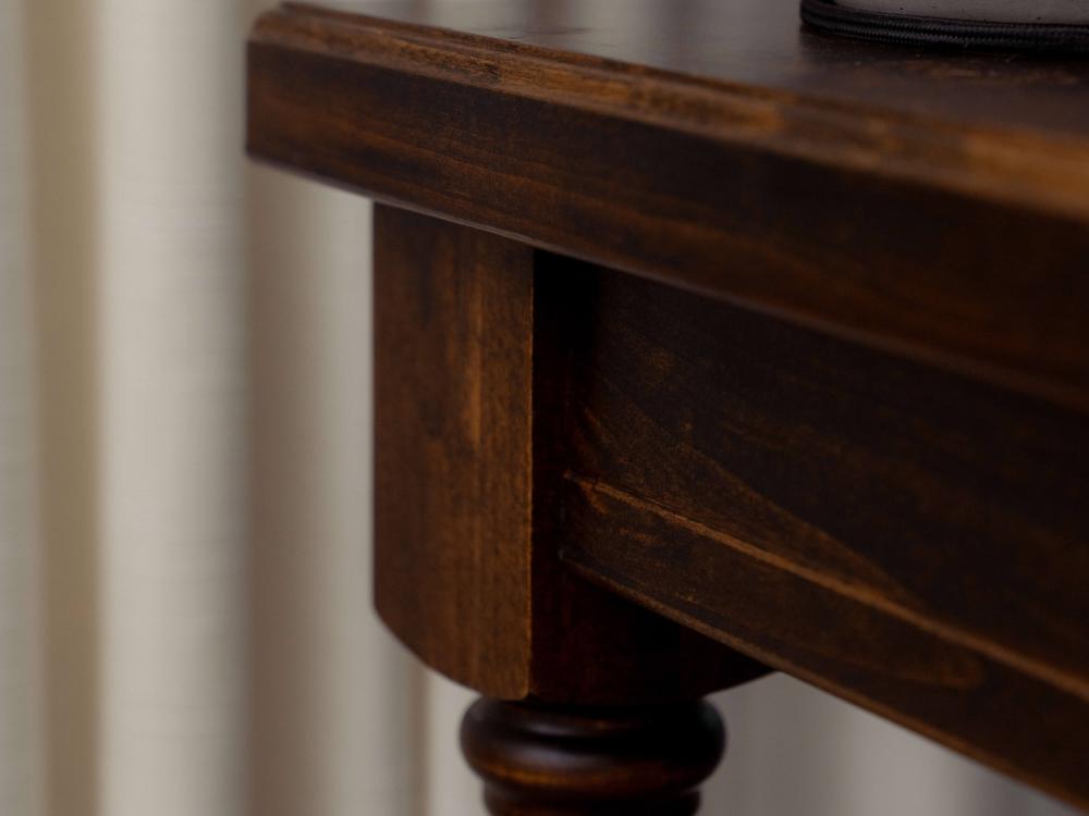 Close-up of the Abigail End Table - Tobacco by James+James, showcasing its dark wooden corner with cylindrical legs. The background is out of focus, highlighting vertical off-white fabric drapes. The tabletop appears smooth and polished.