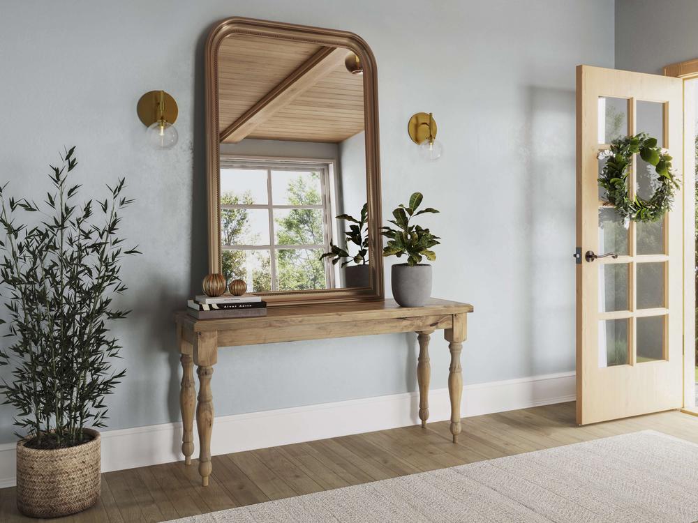 A hallway with an Abigail Sofa Table in Harvest Wheat by James+James, potted plants, a large mirror framed in wood, and wall sconces on either side. An open door with a wreath on it and sunlight streaming in. A woven basket with a tall plant is to the left of the sofa table.