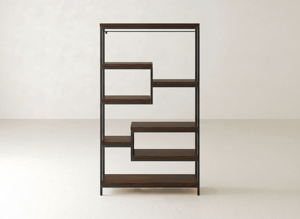 The James+James Arlo Shelf is a modern bookshelf with a geometric, zigzag design. It features multiple solid hardwood shelves framed in industrial steel, creating open spaces of varying heights and widths that are perfect for books or decor. The background consists of plain, light-colored walls and floors.