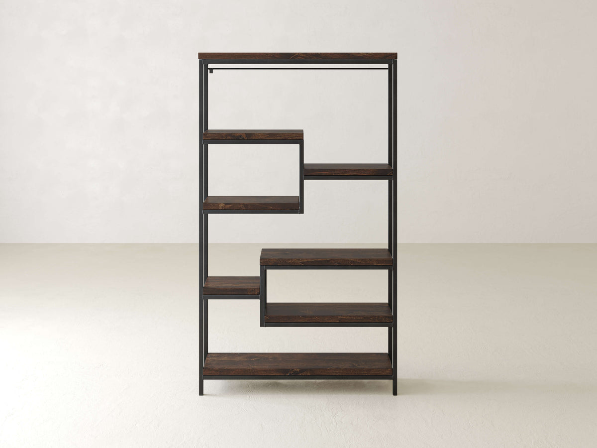 A modern wooden shelf with an asymmetrical, geometric design standing against a plain, light-colored wall. It features five staggered shelves with a metal frame, creating open spaces between each shelf.