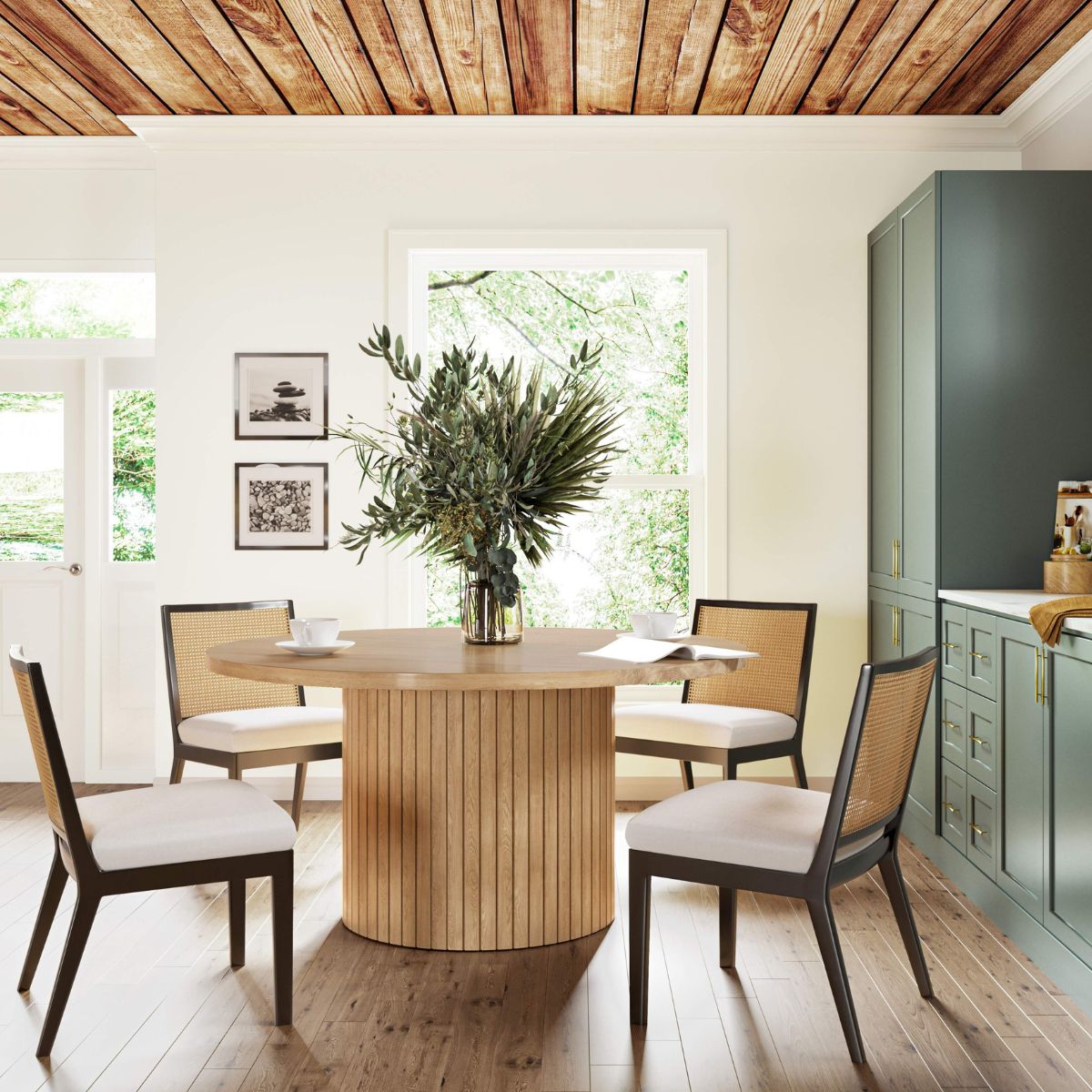 Dining room with a round wooden table and four cushioned chairs. A vase with green foliage sits on the table. The room has a wooden ceiling, large window showing greenery, green cabinets, and framed pictures on a cream wall. Wood flooring throughout.