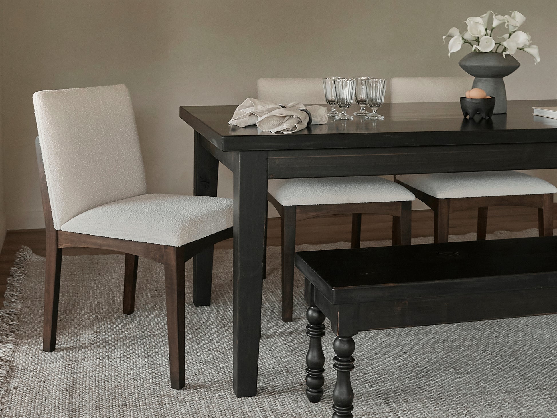 A modern dining room featuring the James+James Ava Parsons Dining Table in Tobacco, cream upholstered chairs, a matching bench, a gray vase with white flowers, glasses, and a linen napkin on a light woven rug.