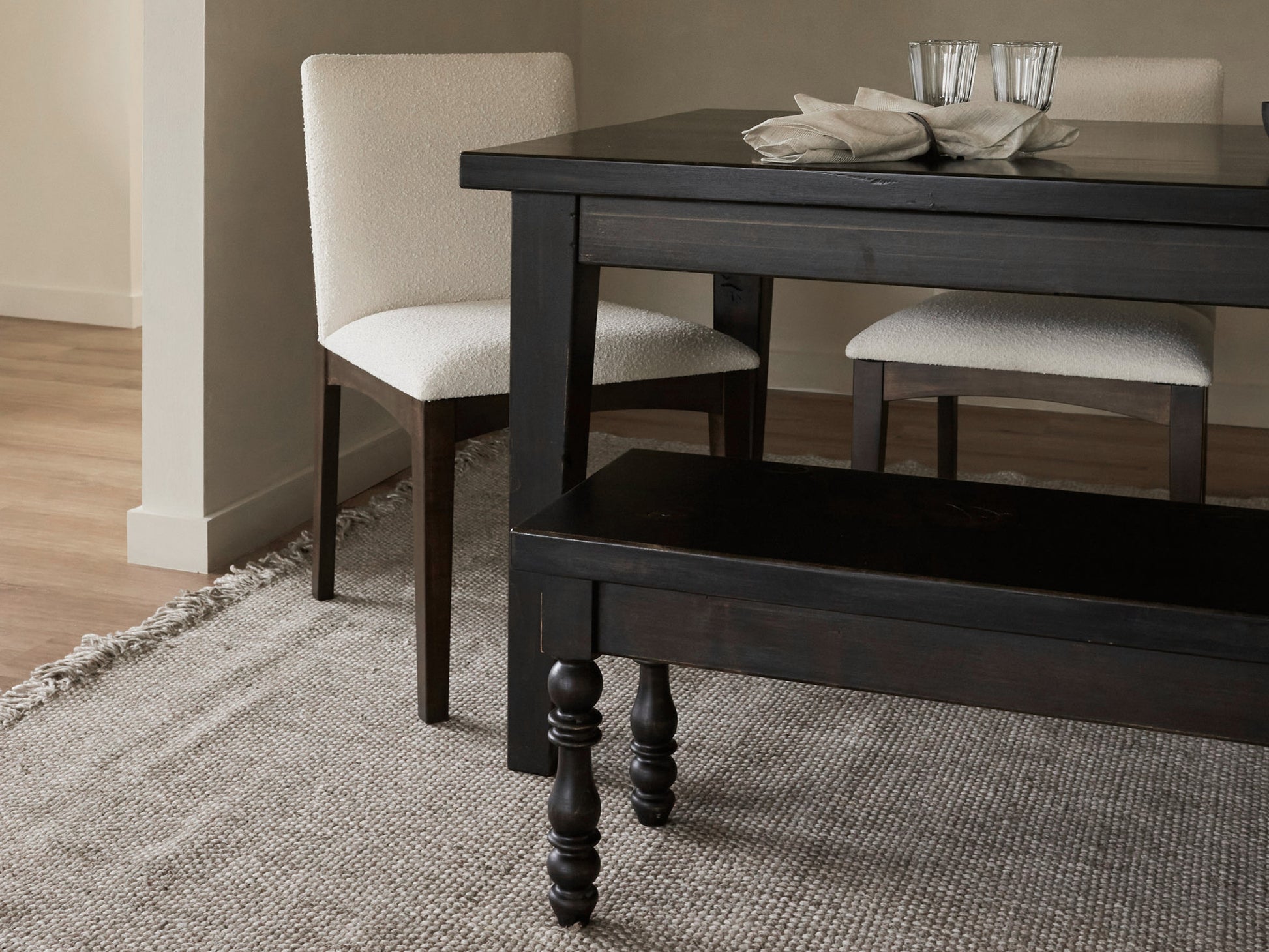 A modern dining area featuring the Ava Parsons Dining Table in Tobacco by James+James, a matching bench, two upholstered white chairs, a grey textured rug, and two glasses with folded napkins on the tabletop.
