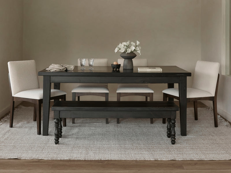 A modern dining room featuring the James+James Ava Parsons Dining Table in Tobacco, four light upholstered chairs, a matching bench, a neutral rug, white flowers in a vase, two glasses, a folded napkin, and a book on the table.