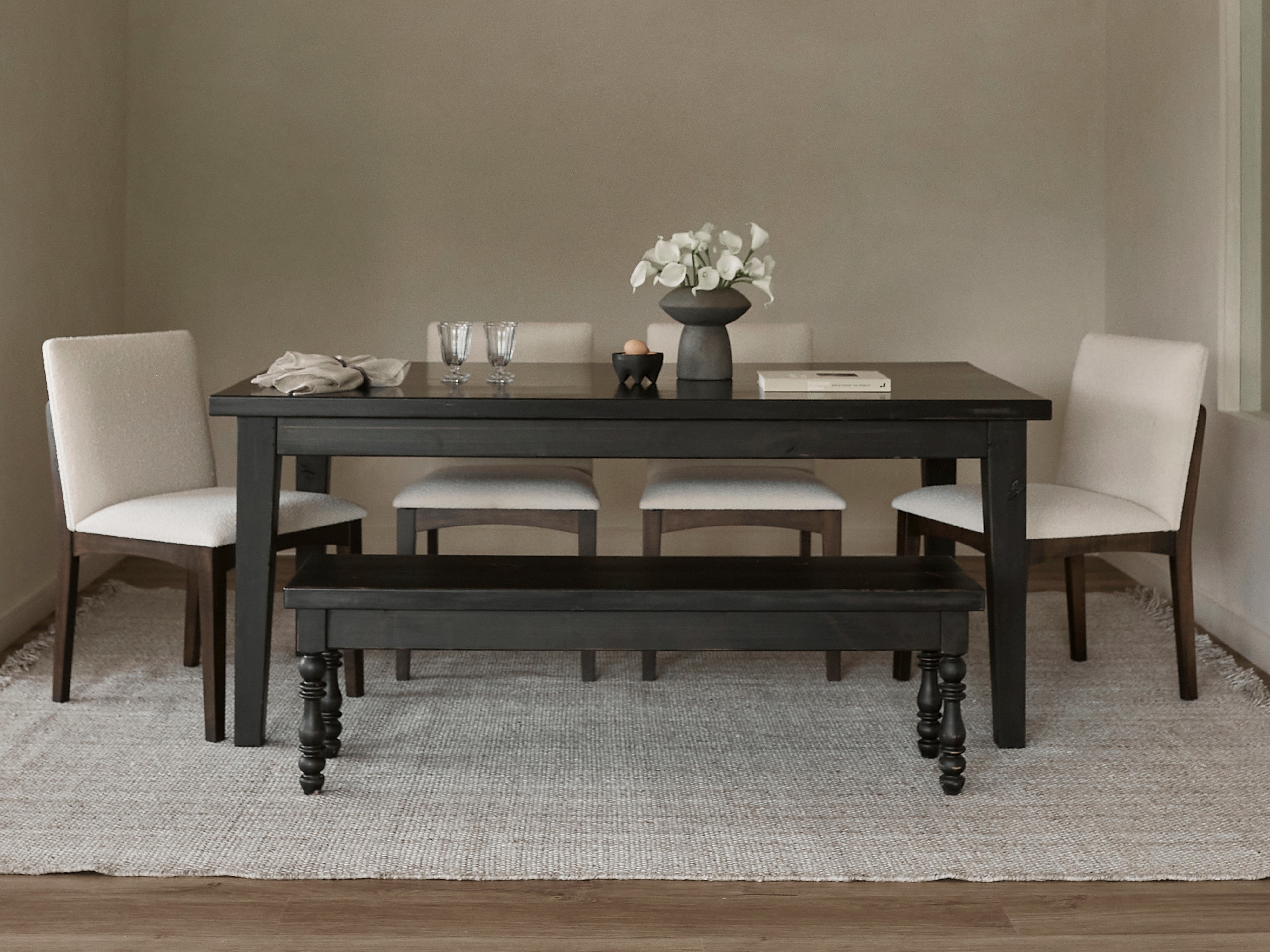 A modern dining room featuring the James+James Ava Parsons Dining Table in Tobacco, four light upholstered chairs, a matching bench, a neutral rug, white flowers in a vase, two glasses, a folded napkin, and a book on the table.
