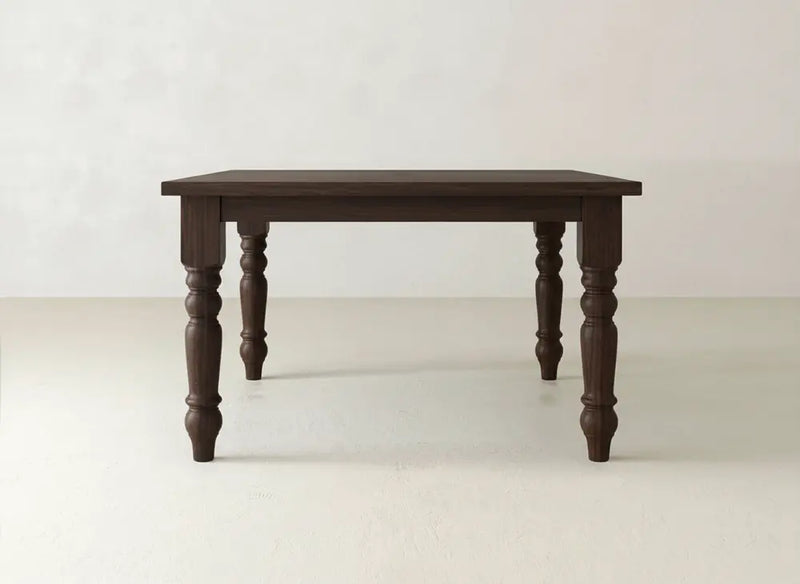 The James & James Fulton Corner Desk in Tuscany features a dark brown rectangular top and four ornate turned legs, set against a plain, light-colored wall and floor.