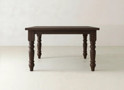 The James & James Fulton Corner Desk in Tuscany features a dark brown rectangular top and four ornate turned legs, set against a plain, light-colored wall and floor.