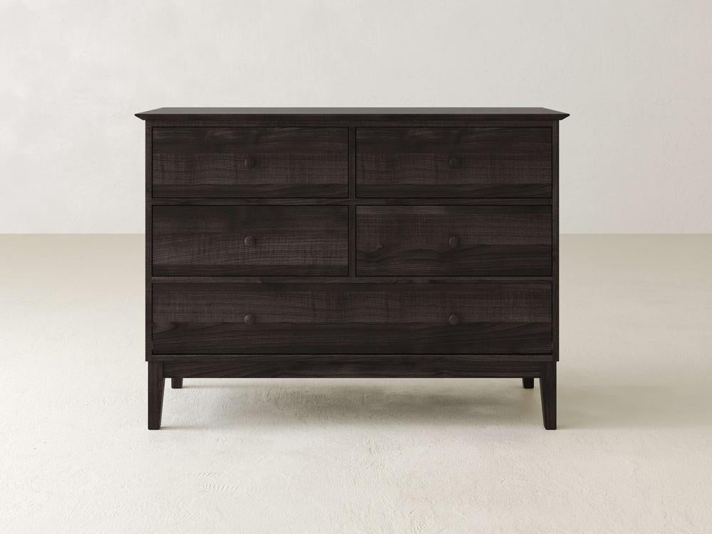 The James & James Bennett 5 Drawer Dresser, made from solid maple, features a sleek black finish with round knobs. Its simple modern design complements an off-white wall and rests elegantly on a light-colored floor.