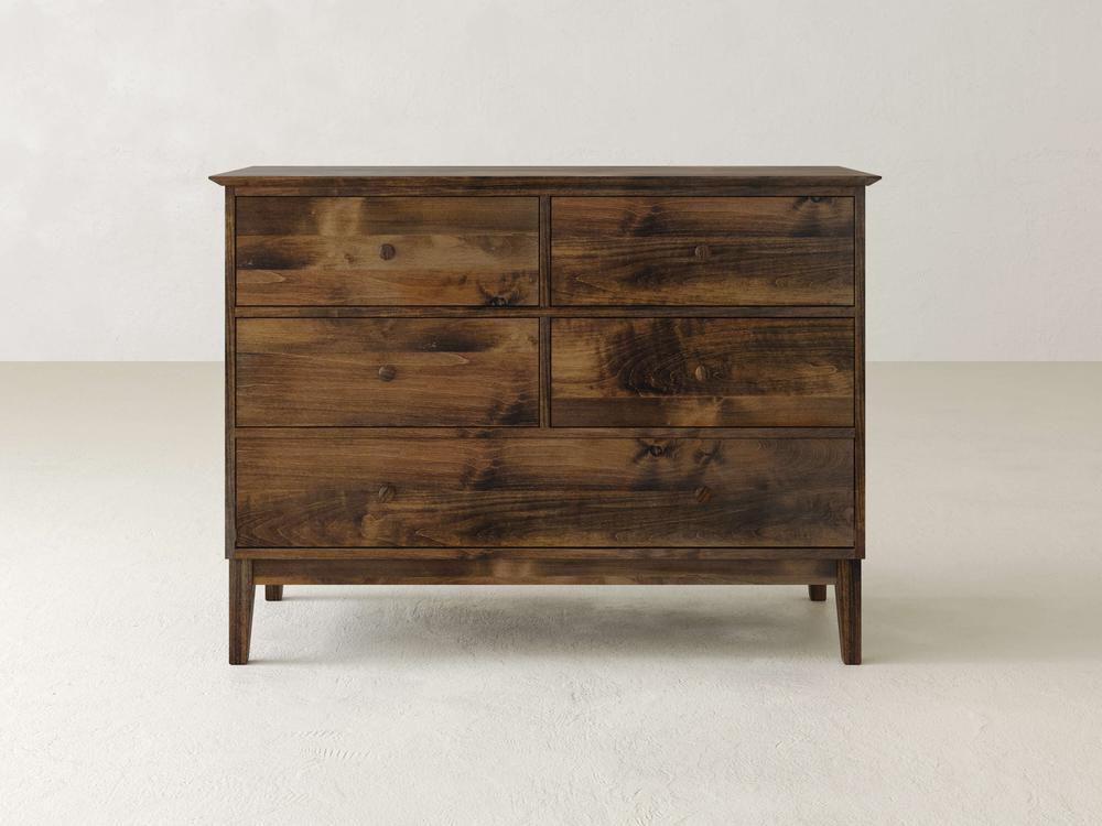 The Bennett 5 Drawer Dresser by James & James is crafted from solid maple with a natural finish, offering a minimalist look. It combines traditional design with clean lines and features five drawers of various sizes against a plain, light-colored background.