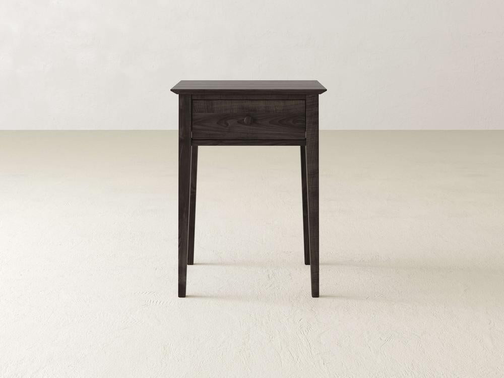 The Bennett 1 Drawer Nightstand by James & James is a dark wood piece made from solid maple, with slender tapered legs, set against a minimalist light background in the bedroom.