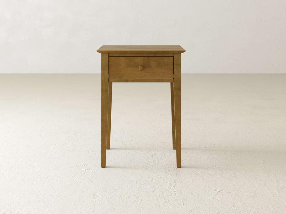 The Bennett 1 Drawer Nightstand by James & James, crafted from solid maple, stands on a light-colored floor. Its minimalist design showcases slender tapered legs, a single drawer, and a smooth surface—ideal for a tranquil bedroom.