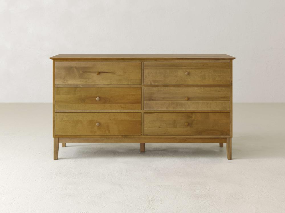 The James & James Bennett 6 Drawer Dresser features a traditional design with simple round knobs. Made from solid maple, it has a light brown finish and stands on four tapered legs, placed against an off-white wall and white floor.