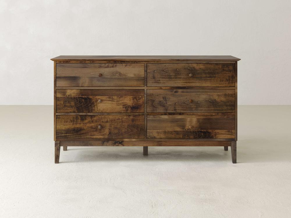 The Bennett 6 Drawer Dresser by James & James is crafted from solid maple wood with smooth-gliding hardware and round handles. It features a natural, rustic finish and stands elegantly on four slightly tapered legs, set against a plain light-colored wall.