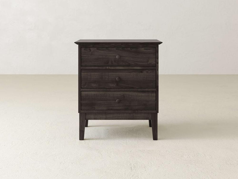 The Bennett 3 Drawer Nightstand by James & James, crafted from solid maple in a dark wooden finish, rests on a light-textured floor against an off-white wall. It features a minimalist traditional design with round knobs on each drawer.